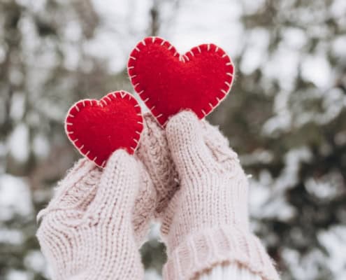 Winter Date-Ideen: Hände in Handschuhen halten zwei rote dekorative Herzen in Winterlandschaft